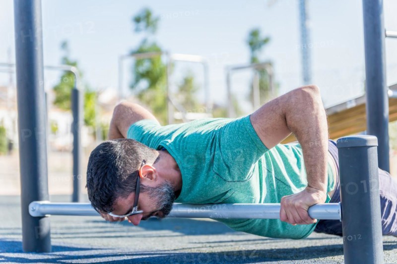 Man seen in profile with beard and sunglasses doing push-ups on a bar on the floor