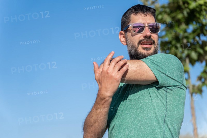 Front view of an attractive bearded man with sunglasses stretching his shoulders in a field with a blue sky in the background