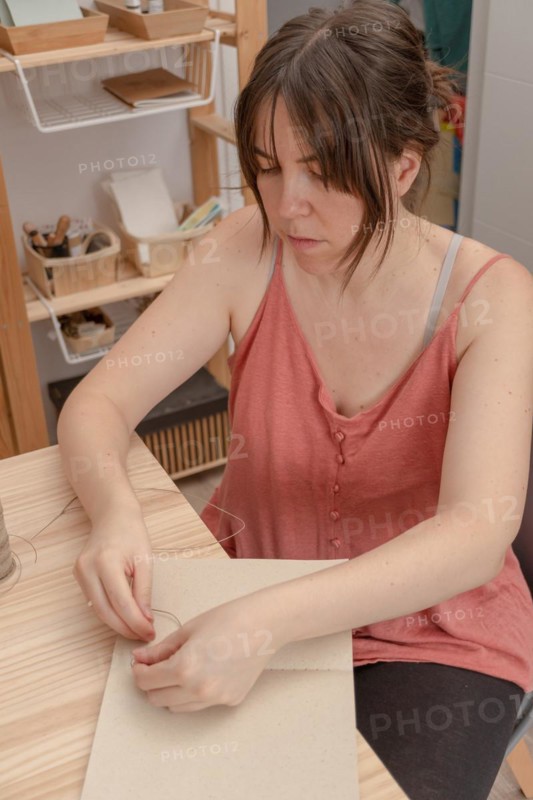 Young brunette girl with long hair craftswoman working on her products in her home workshop sewing notebooks