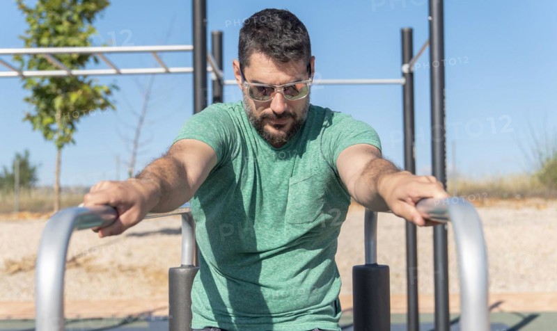 Man with beard and sunglasses training his arms on parallel bars in a park gymn