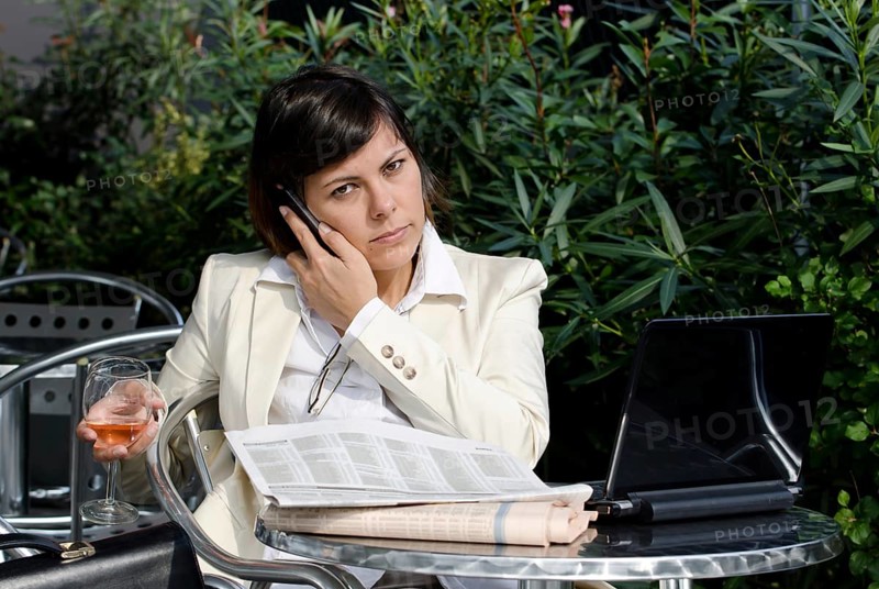 Business Woman Sitting with Her Personal Computer and Financial Newspaper and using Smart Phone on a Table Outdoor