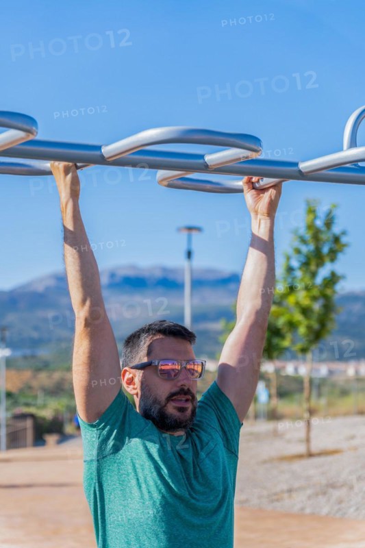 Bearded man with sunglasses seen from the front training his back by doing pull-ups on a barbell assisted by an elastic fitness band in an outdoor gym