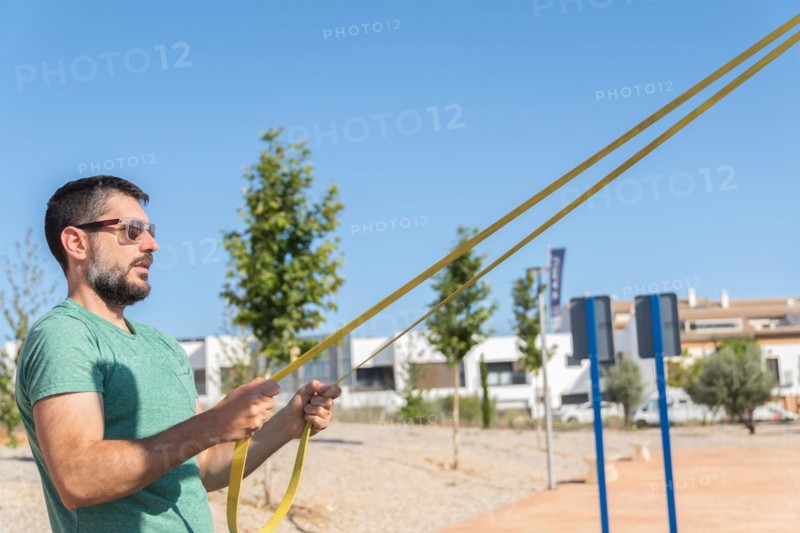 Bearded man with sunglasses seen in profile training his arms with an elastic fitness band in an outdoor gym