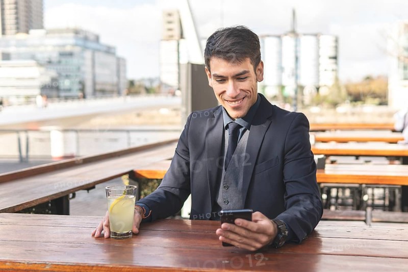 Businessman sitting at an outdoor bar drinking lemonade looking at his phone smiling
