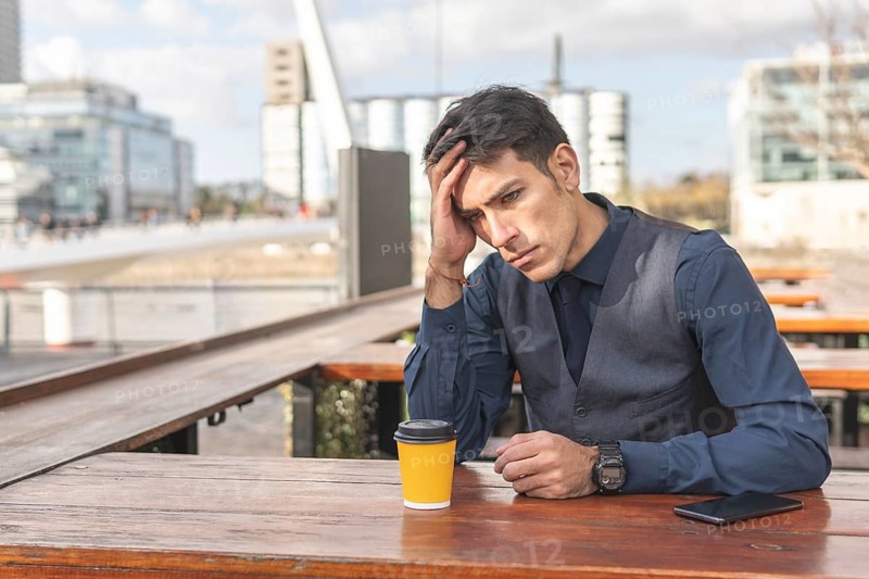 Businessman sitting in an outdoor bar having a coffee clutching his head with a worried expression