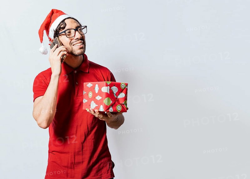 Smiling guy in christmas hat holding christmas gift box and talking on the phone. Christmas man holding gift and calling family on phone. Happy man holding gift and calling family at christmas