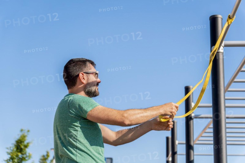 Bearded man with sunglasses seen in profile training with an elastic fitness band in an outdoor gym
