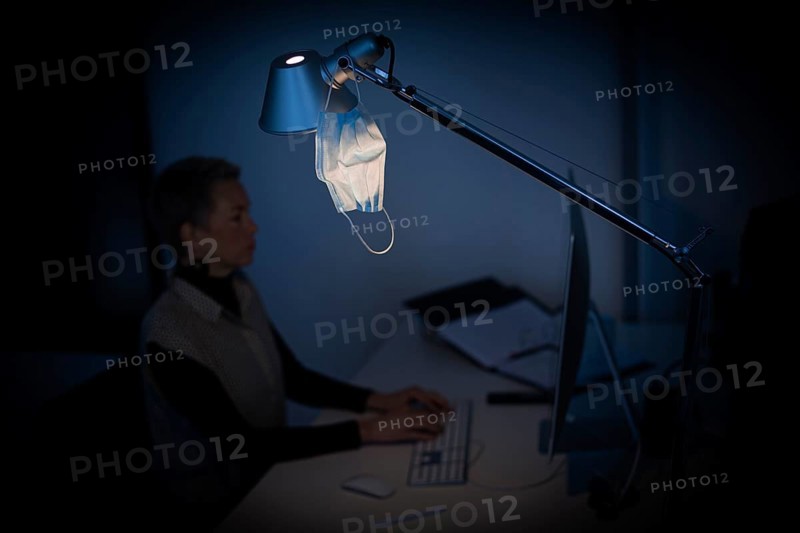 Mask at the workplace. A mask hangs from a lamp above a desk while a woman works on a computer in the background. Berlin