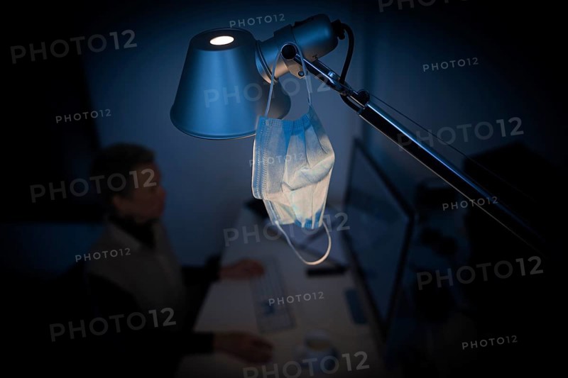 Mask at the workplace. A mask hangs from a lamp above a desk while a woman works on a computer in the background. Berlin