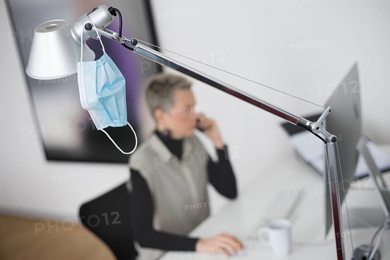 Mask at the workplace. A mask hangs from a lamp above a desk while a woman works on a computer in the background. Berlin