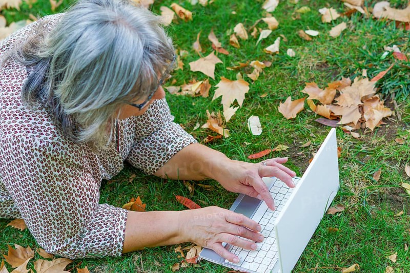 Top view of a white-haired woman working on her laptop computer lying on the grass in a park