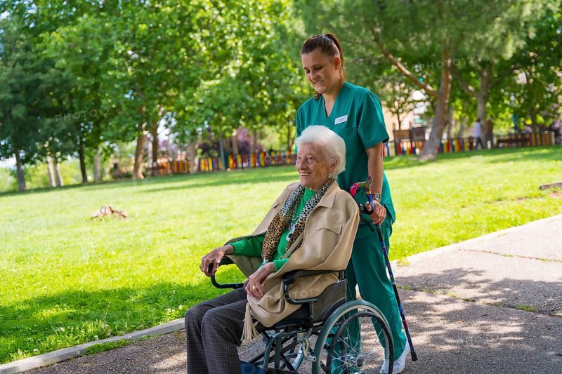 An elderly woman with the nurse on a walk through the garden of a nursing home in a wheelchair next to nature and trees