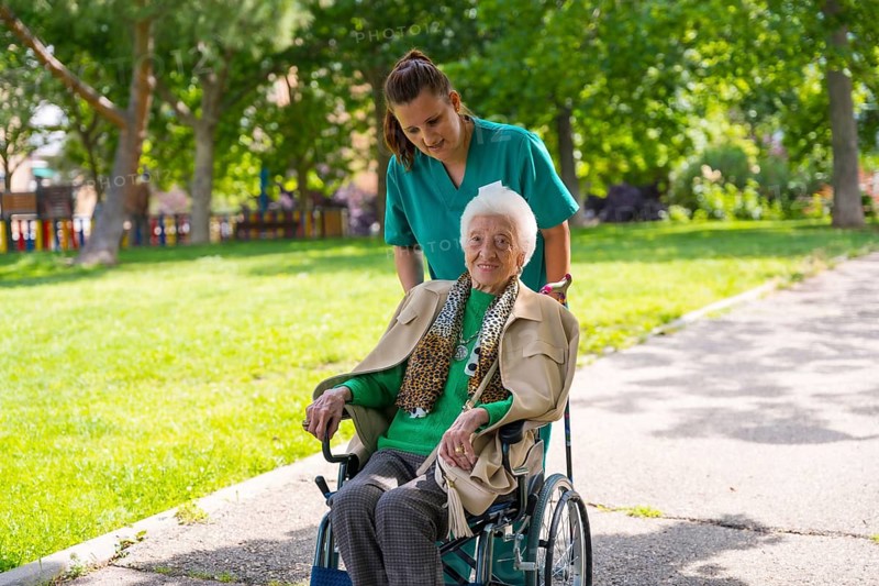 An elderly woman with the nurse on a walk through the garden of a nursing home in a wheelchair next to nature and trees