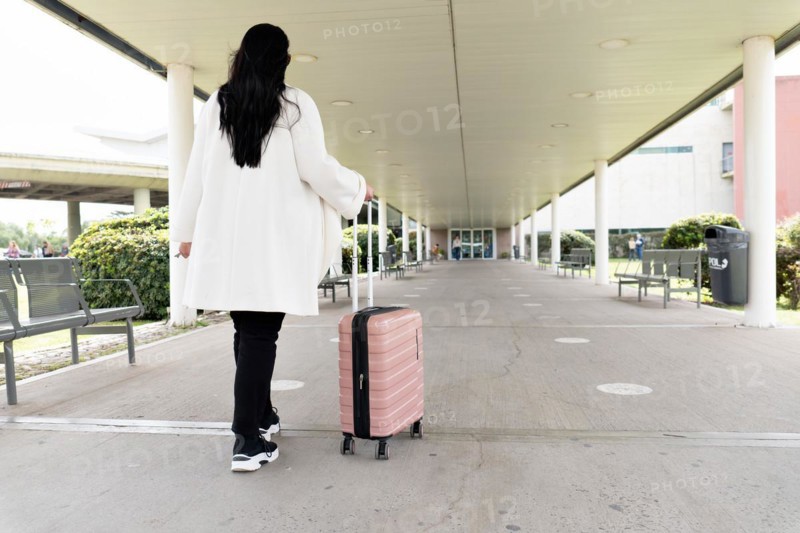 Back shot of woman in sneakers and coat walking towards main entrance of airport and rolling her wheeled suitcase. Travel concept