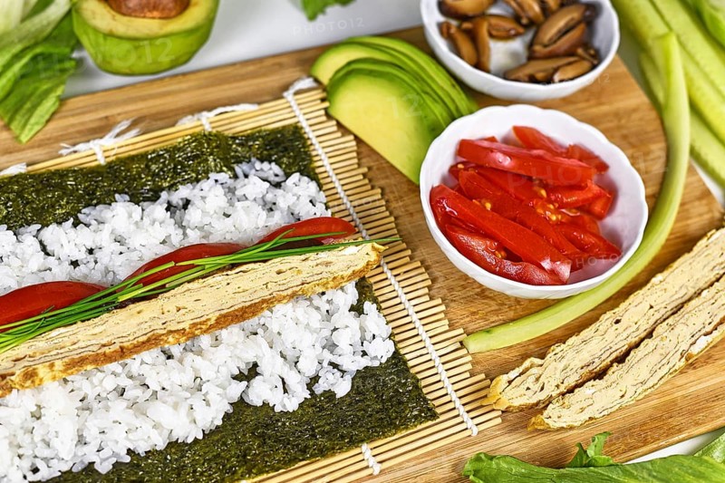 Preparing homemade vegetarian sushi with Nori algea sheet with rice on bamboo mat and ingredients like egg omelet