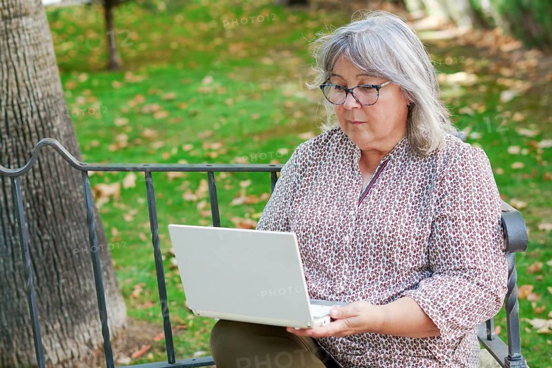 Older white-haired woman working on her laptop computer seated on a park bench