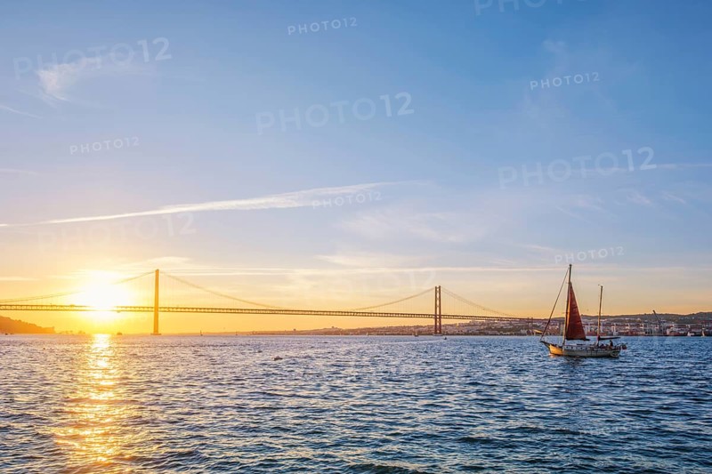 View of 25 de Abril Bridge famous tourist landmark of Lisbon connecting Lisboa and Almada over Tagus river with tourist yacht silhouette at sunset. Lisbon