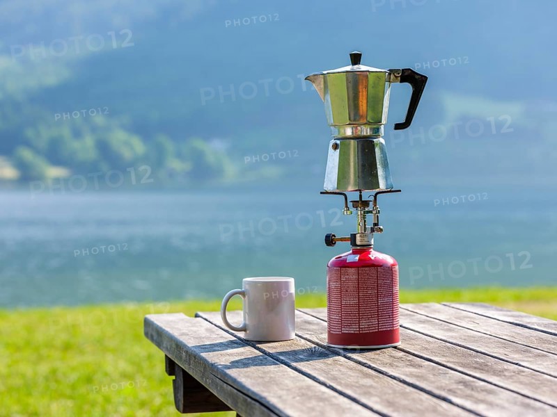 Espresso pot on a gas cooker next to a cup standing on a wooden table at the lake