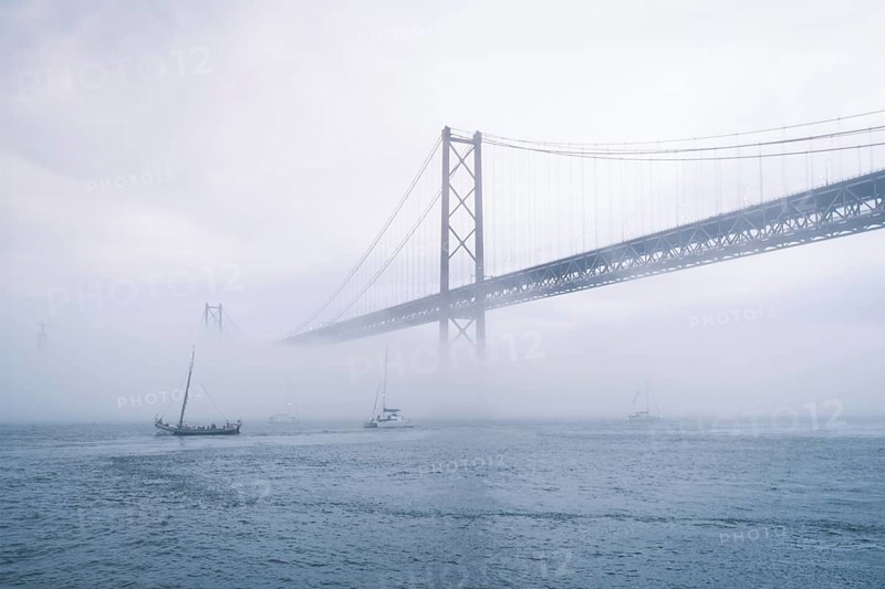 View of 25 de Abril Bridge famous tourist landmark of Lisbon connecting Lisboa and Almada in heavy fog mist wtih yacht boats passing under. Lisbon