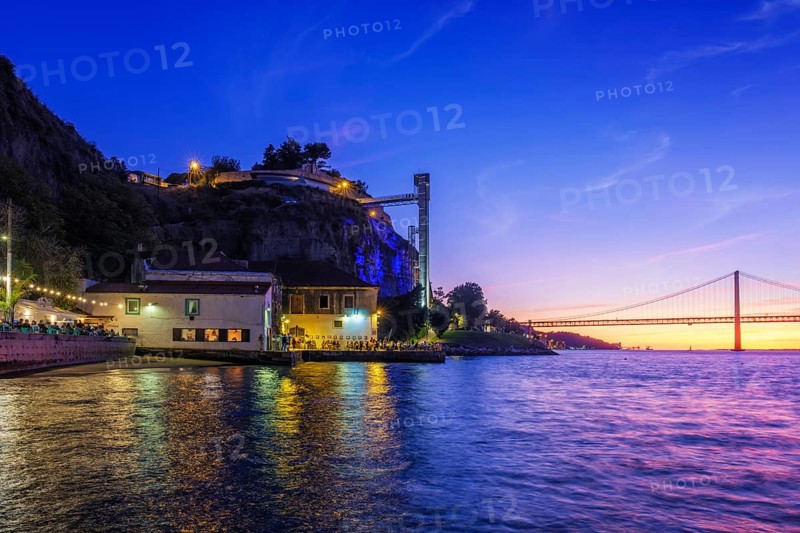 View of 25 de Abril Bridge famous tourist landmark of Lisbon over Tagus river and Boca do Vento Elevator and seaside restaurants with people in the evening. Lisbon