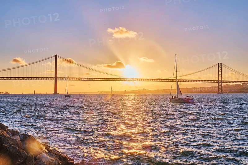 View of 25 de Abril Bridge famous tourist landmark of Lisbon connecting Lisboa and Almada on Setubal Peninsula over Tagus river with tourist yacht silhouette at sunset. Lisbon