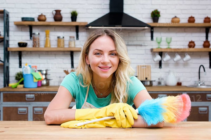 Smiling housekeeper leaning kitchen counter holding feather duster looking camera