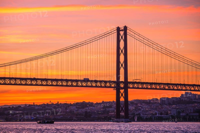 View of 25 de Abril Bridge famous tourist landmark of Lisbon connecting Lisboa and Almada over Tagus river with tourist boat silhouette at sunset. Lisbon