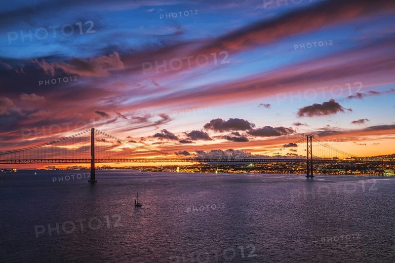 View of 25 de Abril Bridge famous tourist landmark of Lisbon connecting Lisboa and Almada on Setubal Peninsula over Tagus river in the evening twilight with boats. Lisbon