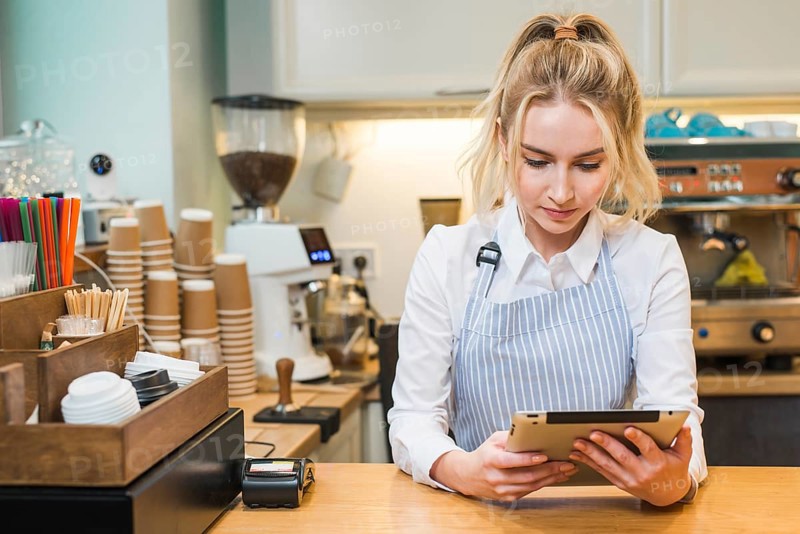 Blonde young woman standing coffee shop counter looking digital tablet