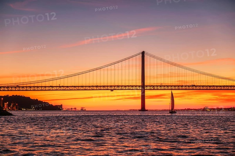 View of 25 de Abril Bridge famous tourist landmark of Lisbon connecting Lisboa and Almada over Tagus river with tourist yacht silhouette at sunset. Lisbon