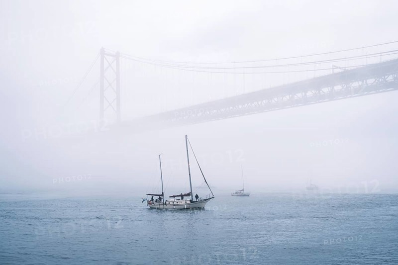 View of 25 de Abril Bridge famous tourist landmark of Lisbon connecting Lisboa and Almada in heavy fog mist wtih yacht boats passing under. Lisbon