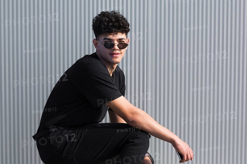 Young ethnic curly man sunglasses black clothes against grey wall