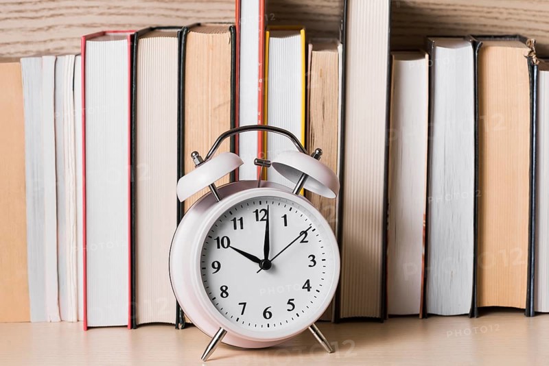 White alarm clock showing 10 o clock front bookshelf wooden desk