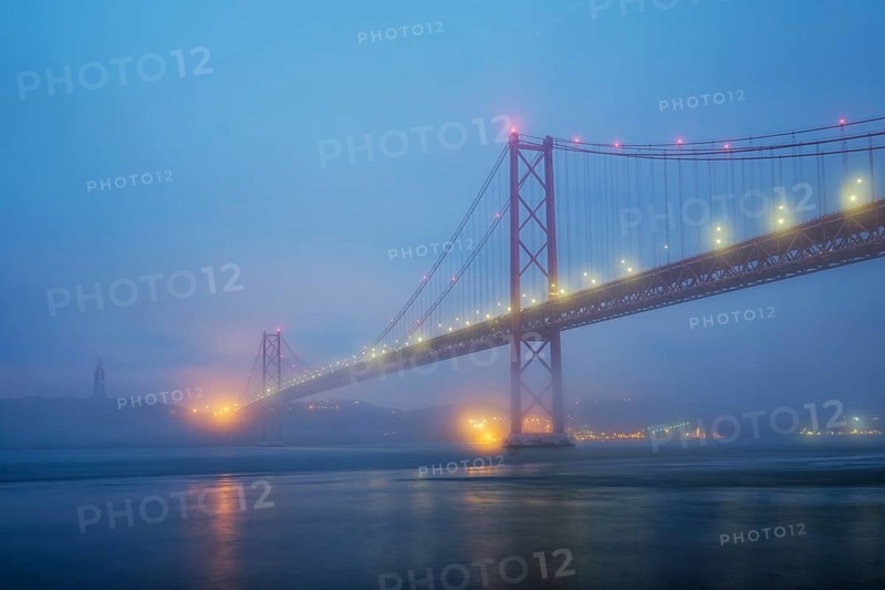 View of 25 de Abril Bridge famous tourist landmark of Lisbon connecting Lisboa and Almada in heavy fog mist wtih yacht boats passing under. Lisbon