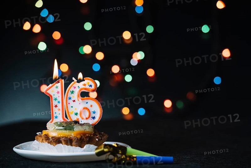 Number 16 birthday lighted candle slice tart with party horn blower against illuminated bokeh backdrop