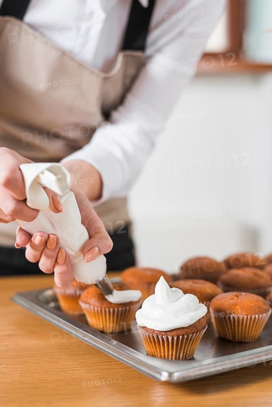 Young woman decorating cupcakes with white whipped cream by squeezing confectionery bag