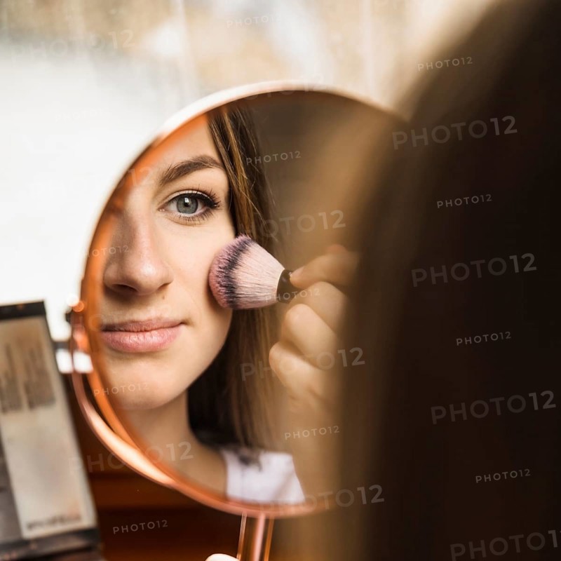 Hand mirror with reflection woman applying blusher her face