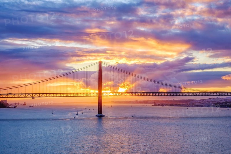 View of 25 de Abril Bridge famous tourist landmark of Lisbon connecting Lisboa and Almada on Setubal Peninsula over Tagus river with tourist yacht boats on sunset. Lisbon