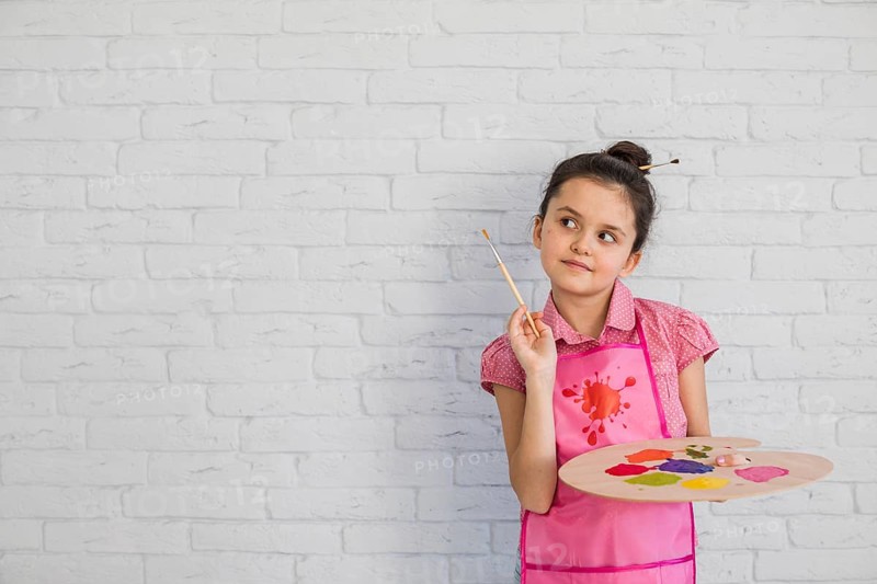 Portrait girl with paintbrush palette standing against white wall