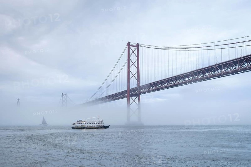 View of 25 de Abril Bridge famous tourist landmark of Lisbon connecting Lisboa and Almada in heavy fog mist wtih yacht boats passing under. Lisbon