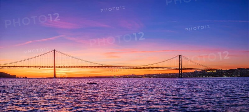 Panorama of 25 de Abril Bridge famous tourist landmark of Lisbon connecting Lisboa and Almada over Tagus river with tourist yacht silhouette at sunset. Lisbon