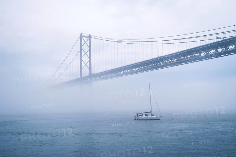 View of 25 de Abril Bridge famous tourist landmark of Lisbon connecting Lisboa and Almada in heavy fog mist wtih yacht boats passing under. Lisbon