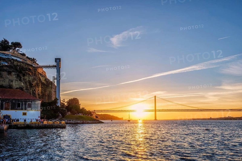 View of 25 de Abril Bridge famous tourist landmark of Lisbon over Tagus river and Boca do Vento Elevator and seaside restaurants with people at the sunset. Lisbon