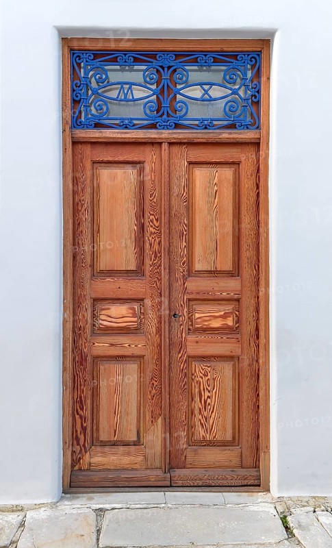 Old vintage wooden house doors in light natural color decorated with blue forge-smithing metal ornament with monograms of capital letters M and X. Whitewashed walls