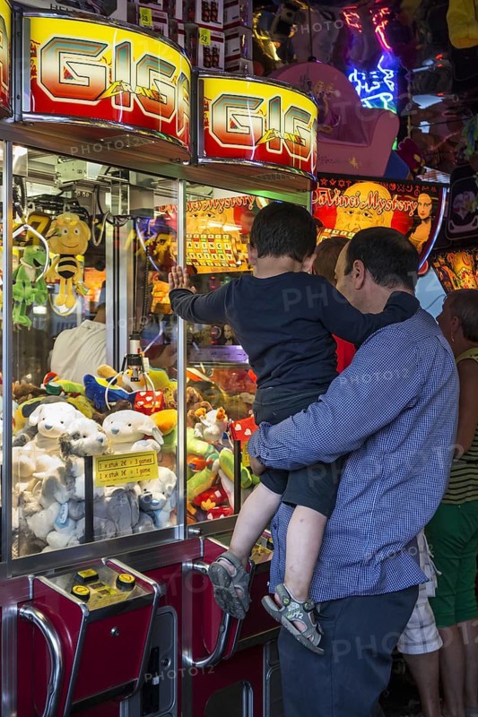 Father with son looking at claw crane game machine filled with toys in amusement arcade at travelling funfair