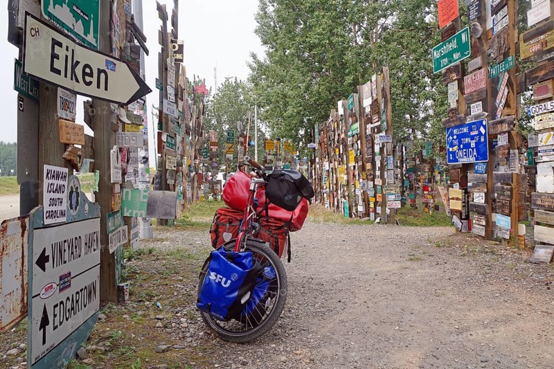 Various traffic and place signs face a bicycle loaded with luggage