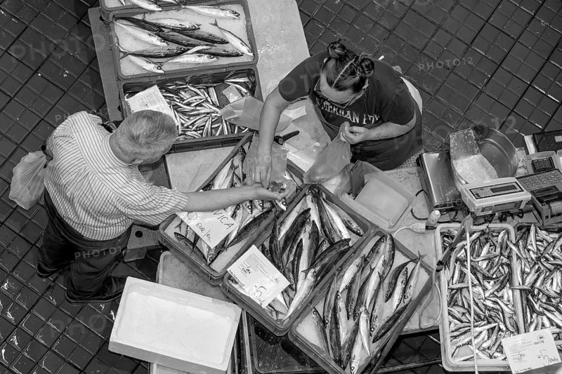 Black and white bird's eye view photo of a stall full of fish with the seller and a buyer