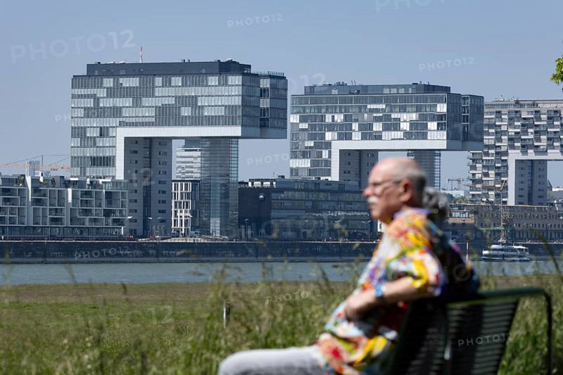 Senior couple sitting on a bench on the banks of the Rhine in front of the crane houses in Cologne's Rheinauhafen harbour on the other bank