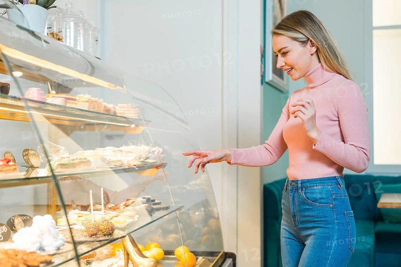 Smiling young woman choosing cake standing front glass cabinet