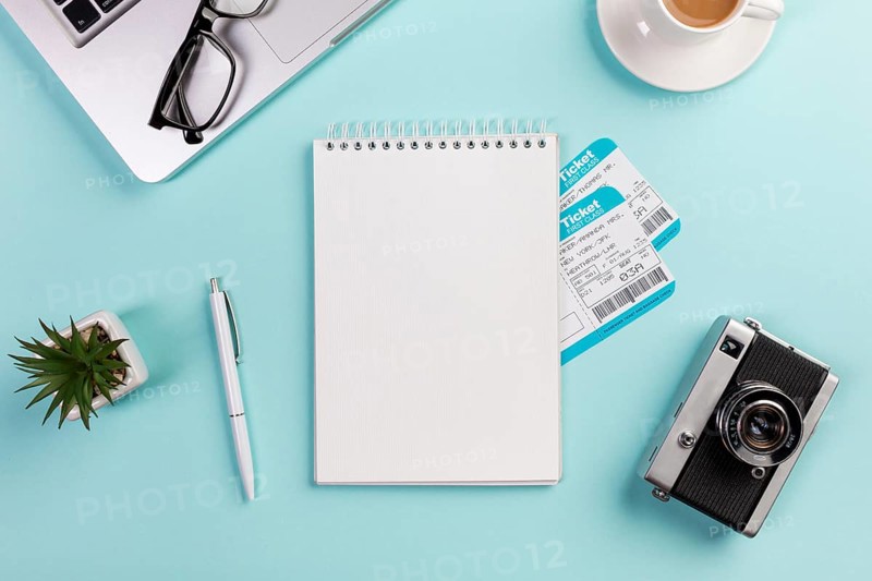 Blank spiral notepad with air tickets surrounded with laptop eyeglasses pen camera coffee cup blue desk